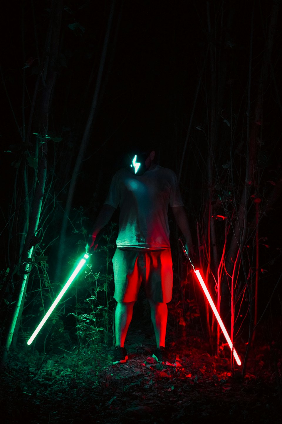 Masked person holding glowing swords in an overgrown forest at night.